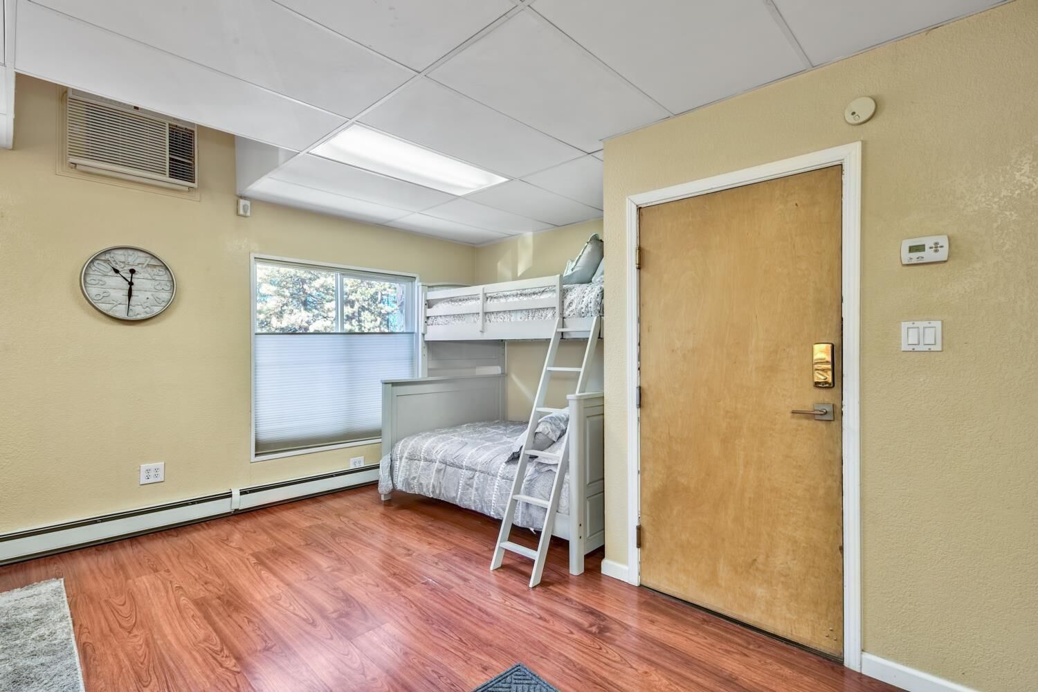 A bedroom with a bunk bed, a clock on the wall, a window with blinds, and a door with a thermostat.