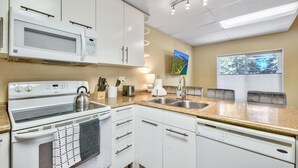 Une cuisine moderne avec des meubles blancs, un four à micro-ondes, une cuisinière et un évier. Il y a une fenêtre avec des stores et un tableau au mur.