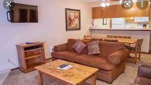 Ein Wohnzimmer mit braunem Sofa, einem Holztisch, einem Fernseher und einer Küchenzeile im Hintergrund.