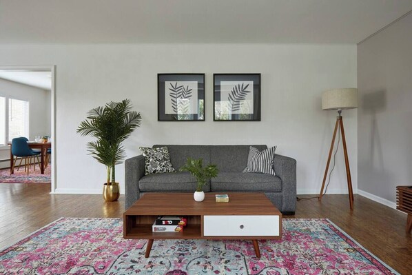 A living room with a grey sofa, two framed leaf artworks, a coffee table with books, a floor lamp, and a large potted plant. - Ballard Hot Tub Retreat + Fenced Yard + Walk to Cafes Dining & Breweries (Seattle)
