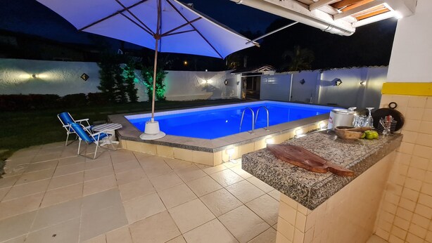 A swimming pool with a blue interior, surrounded by a tiled patio area with a sun umbrella, a lounge chair, and a small kitchen counter with various items.