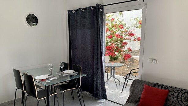 Zona pranzo moderna con tavolo e sedie in vetro, una poltrona con cuscino decorato e vista su un giardino con fiori rossi attraverso una porta scorrevole.