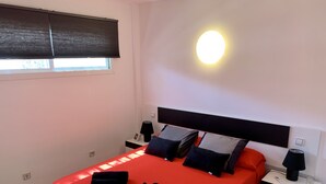 Camera da letto con copriletto rosso, cuscini neri e grigi, un comodino con una lampada e una luce a parete.