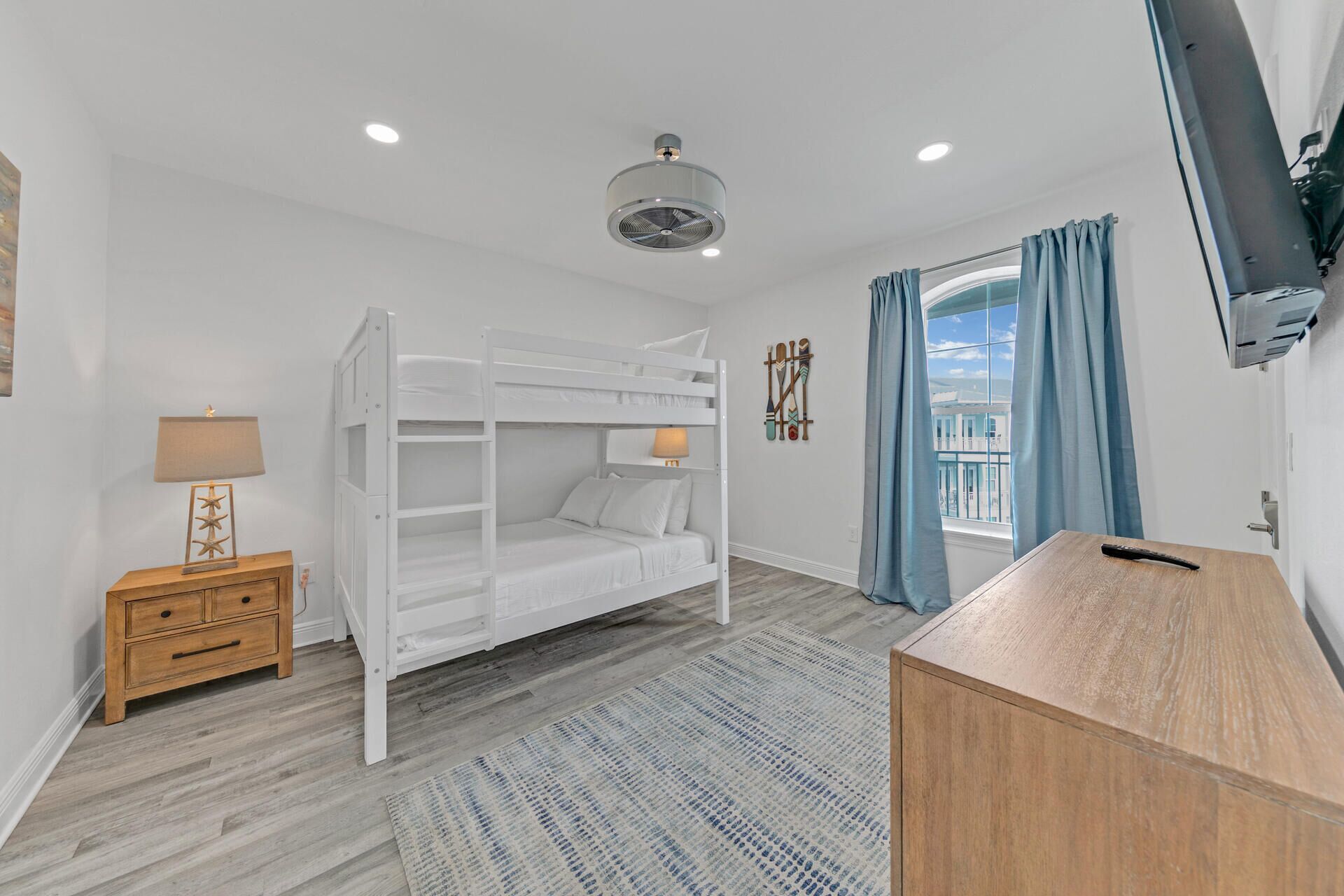 A bunk bed room with a wooden nightstand, a desk, a wall-mounted TV, and a window with curtains.