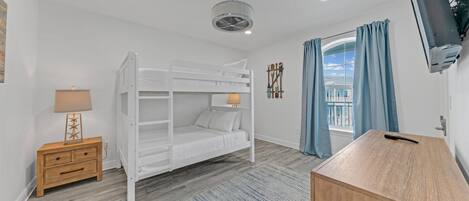 A bunk bed room with a wooden nightstand, a desk, a wall-mounted TV, and a window with curtains.