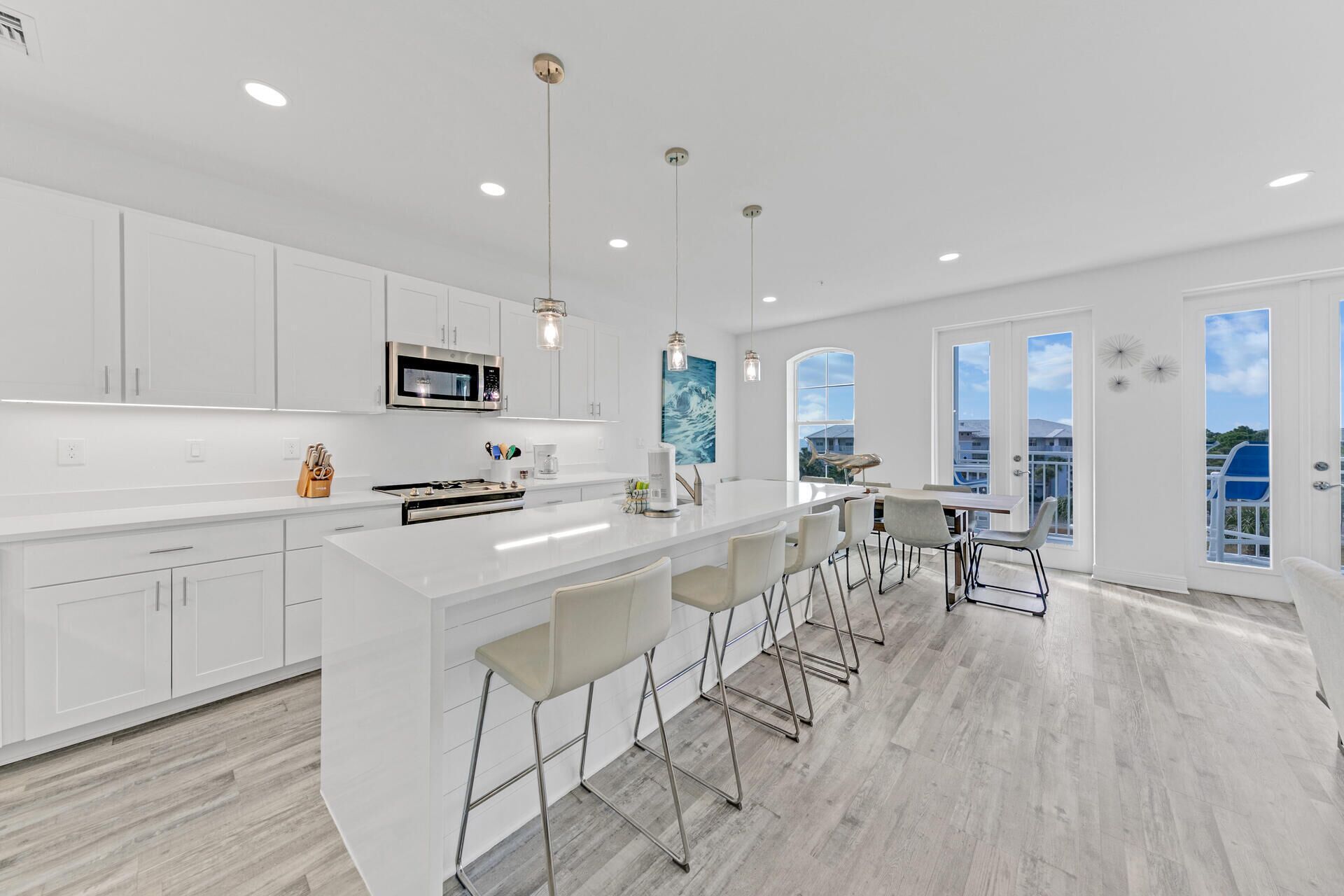 Una cocina moderna con armarios blancos, una isla grande con taburetes y vistas al exterior gracias a amplios ventanales.