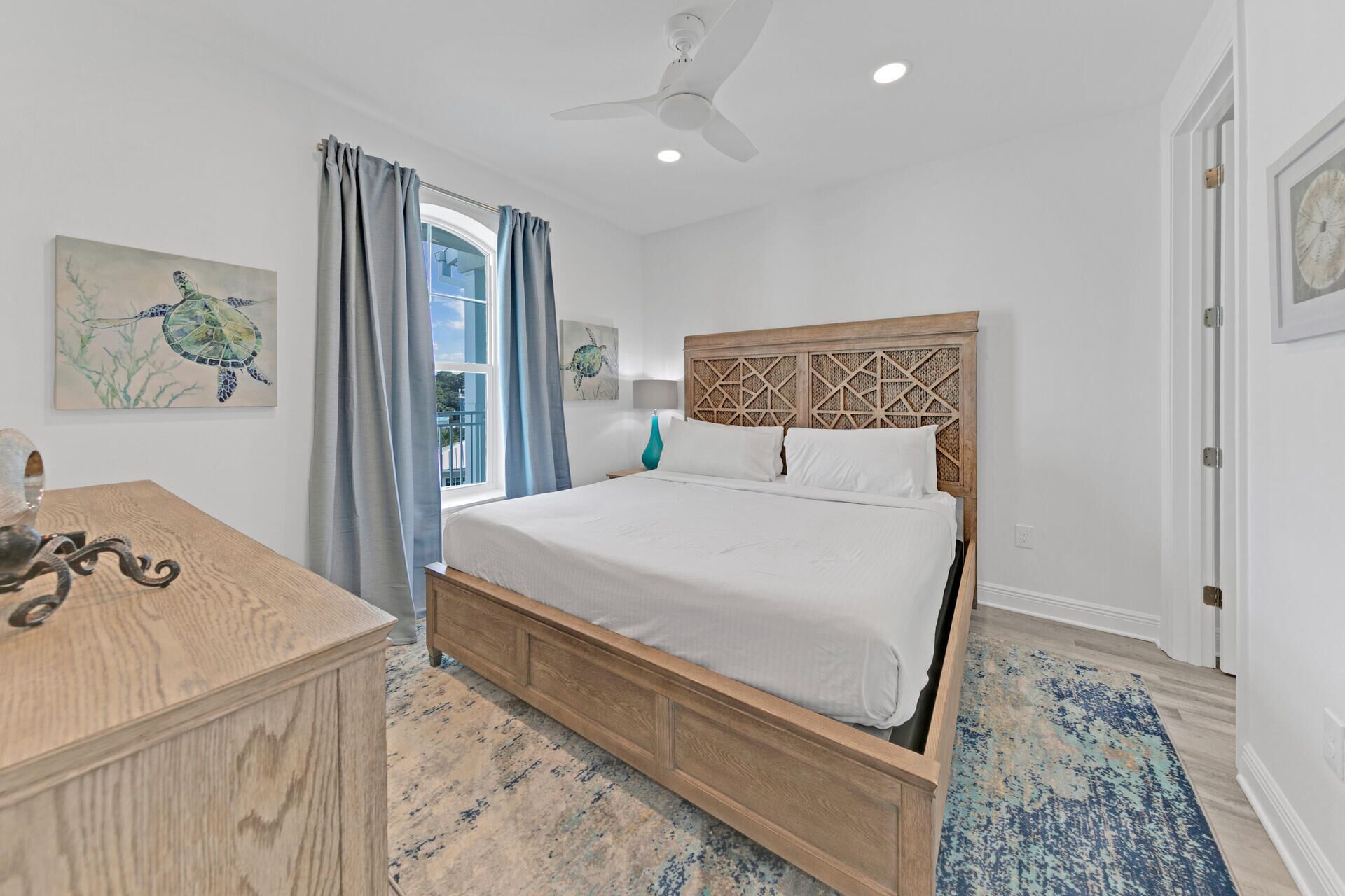 A bedroom with a wooden bed, a ceiling fan, and a view of the outdoors through a window with blue curtains.