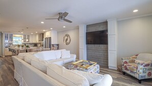 Ein modernes Wohnzimmer mit einer Couch, einem Couchtisch und einem gemusterten Sessel. Im Hintergrund ist eine Küche erkennbar, mit einem großen Fernseher an der Wand.