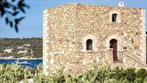 Un edificio de piedra con ventanas de arco y un balcón, rodeado de vegetación y con vistas a un cuerpo de agua.