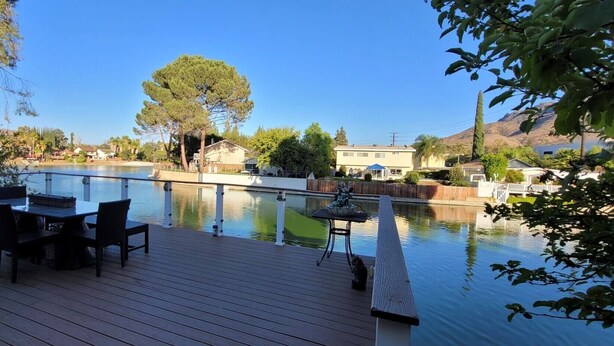 A wooden deck with a table and chairs overlooking a calm body of water, surrounded by trees and houses. - Lakefront 3 Bedroom house with breath taking views (Agoura Hills)