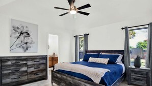 Ein Schlafzimmer mit Bett, Schminktisch, Nachttisch, Deckenventilator und einem Fenster mit Vorhängen.