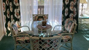 Un comedor con una mesa de vidrio rodeada de sillas con estampado floral, un ventilador de techo y cortinas translúcidas con diseños florales.