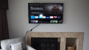 Ein Wohnzimmer mit Flachbildfernseher über einem Kamin, ein grauer Sessel mit gestreiftem Kissen und ein gewebter Korb in der Kaminvertiefung.