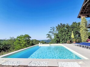 A rectangular swimming pool with clear blue water, surrounded by a stone deck and a lush green landscape.