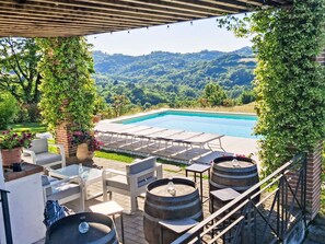 An outdoor pool area with lounge chairs and tables, surrounded by greenery and hills. - Sant'Evasio by Interhome (Ovada)