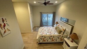 A bedroom with a bed, a nightstand, a ceiling fan, a window with a view, and a door leading to a balcony.