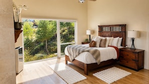 A bedroom with a large bed, bedside tables, a view of greenery outside, and a sliding glass door. - Colorado Mountain Villa (Morrison)