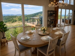 A dining area with a round table set for six, surrounded by white chairs, with a view of a forested landscape through large windows. - Colorado Mountain Villa (Morrison)