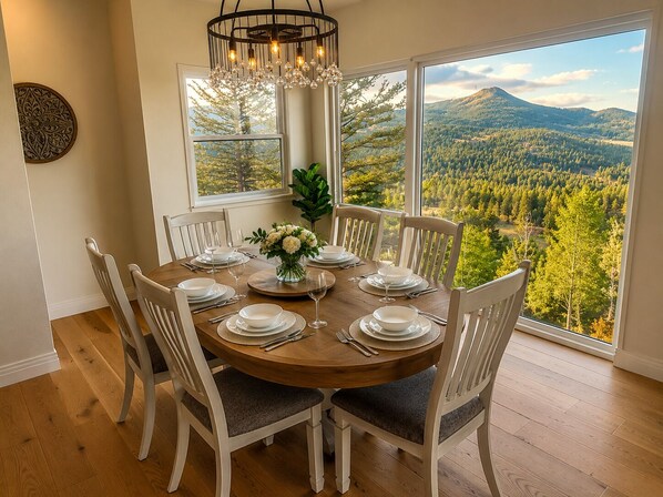 A dining area with a round table set for six, surrounded by white chairs with grey cushions. A chandelier hangs above the table, and a vase with white flowers is placed in the center. Large windows offer a view of a forested landscape with mountains in the distance. - Colorado Mountain Villa (Morrison)