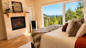A bedroom with a fireplace, a large window with a view of trees, and a bed with pillows. - Colorado Mountain Villa (Morrison)