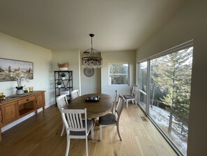 A dining area with a wooden table, white chairs, a chandelier, and a view of trees through large windows.