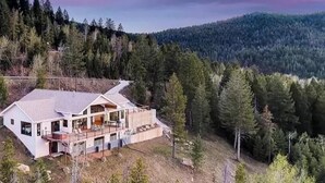 A modern two-story house with a large balcony, surrounded by dense forest and hills.