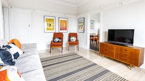 A modern living room with a sofa, two armchairs, a TV on a wooden cabinet, and framed artwork on the walls. - Stylish Bondi Beach pad moments from sand & surf (Bondi Beach)