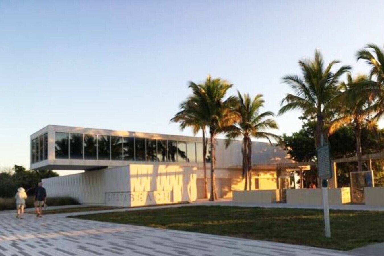 Um edifício moderno com telhado plano e grandes janelas, rodeado por palmeiras e um caminho pavimentado.