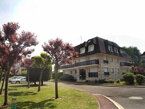 Um edifício residencial de vários andares com uma varanda no segundo pavimento, rodeado por árvores e uma área ajardinada.