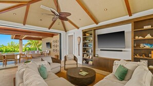 A spacious living room with a large sofa set, a coffee table, and a television mounted on the wall. There is a bookshelf with decorative items and a ceiling fan. - Maluhia Makai: Tropical Villa (Koloa)