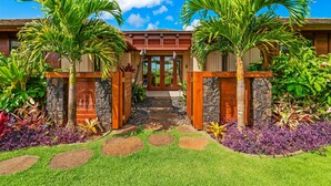 A house with a stone pathway leading to a wooden door, surrounded by lush greenery and palm trees. - Maluhia Makai: Tropical Villa (Koloa)