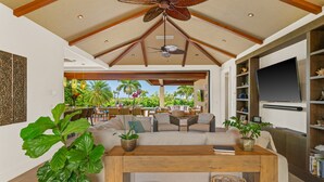 A spacious living room with a high ceiling, a wooden coffee table, and a large sofa. There is a TV mounted on the wall and a view of the outdoors through an open door. - Maluhia Makai: Tropical Villa (Koloa)