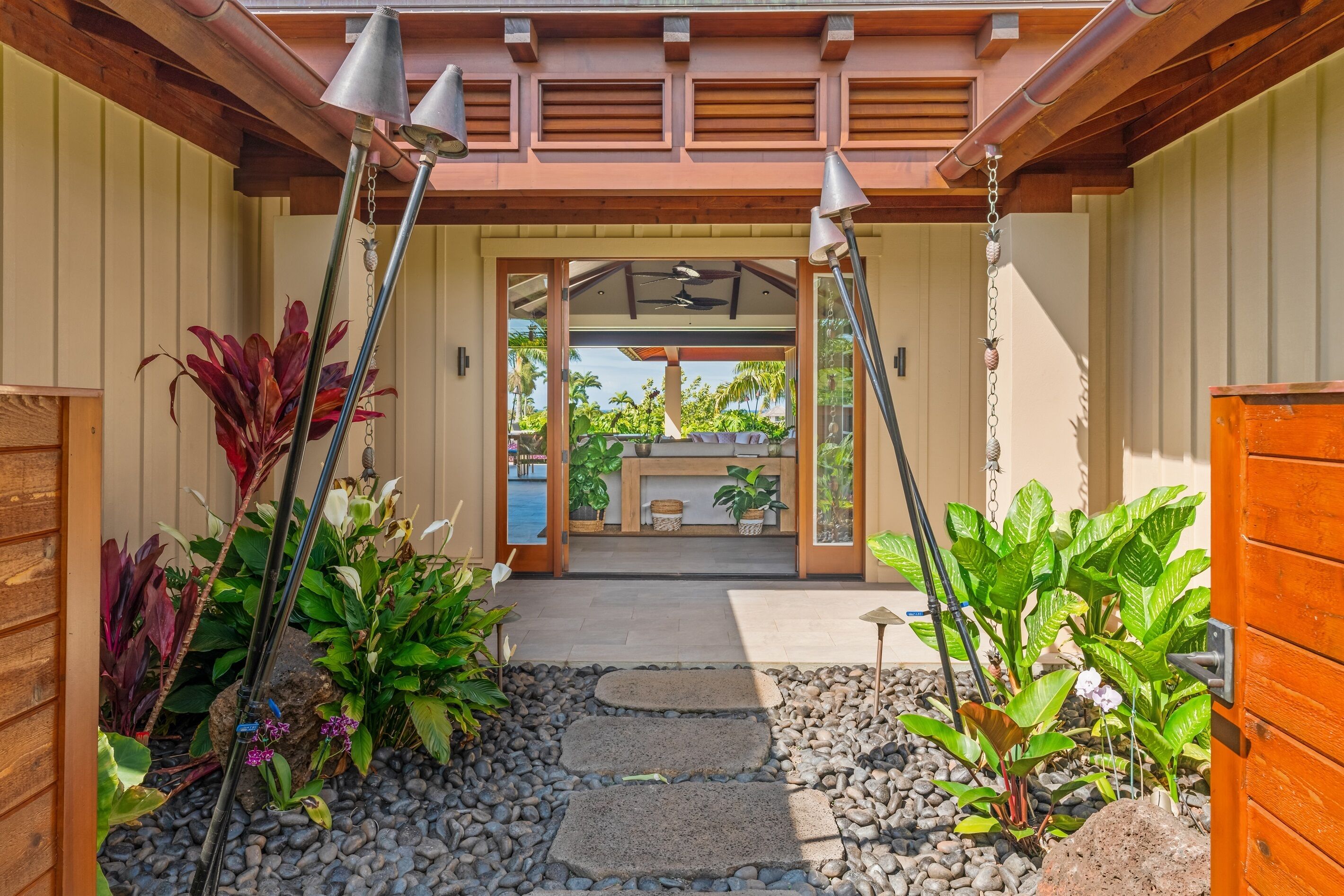 A pathway leading to a covered entrance with a ceiling fan, surrounded by potted plants and a stone-tiled area.
