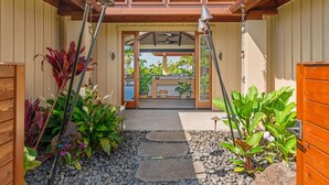 A pathway leading to a covered entrance with a ceiling fan, surrounded by potted plants and a stone-tiled area. - Maluhia Makai: Tropical Villa (Koloa)