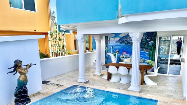 Una piscina con una estatua, un comedor con sillas blancas y un mural de paisaje costero.