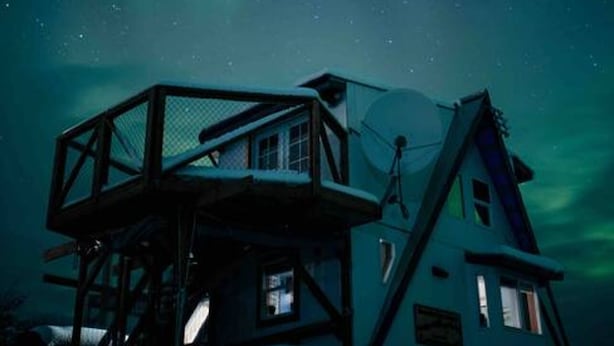 A house with a viewing platform and a satellite dish, set against a night sky with visible stars.