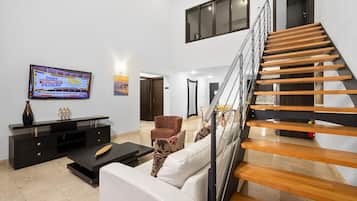 Ein modernes Wohnzimmer mit einer schwarzen Unterhaltungsecke, einem weißen Sofa, einem gemusterten Sessel und einer Holztreppe.
