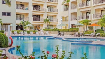 Ein Hotelpoolbereich mit mehreren Liegestühlen und Sonnenschirmen, umgeben von einem mehrstöckigen Gebäude mit Balkonen.