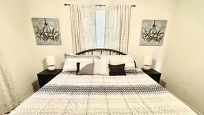 Ein Bett mit gemustertem Überwurf, zwei Nachttische mit Lampen sowie zwei floral gestaltete Wandbilder.