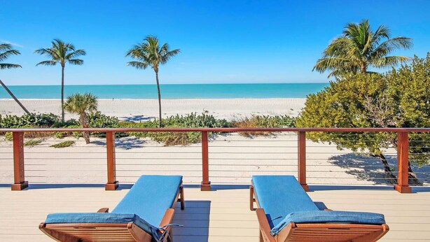 A wooden deck with two lounge chairs facing a beach with palm trees and a clear blue sky.