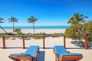 Eine Holzterrasse mit zwei Liegestühlen vor einem Strand mit Palmen und einem klaren, blauen Himmel.