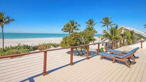 A wooden deck with lounge chairs overlooking a beach with palm trees and a clear blue sky.
