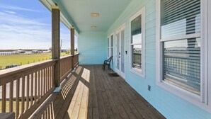 Une véranda couverte avec une balustrade en bois, une vue sur un champ et une rangée de fenêtres équipées de stores.