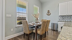 Un coin repas avec une table dressée pour quatre personnes, entouré de chaises en osier, un vase contenant des plumes d’herbe des pampas et une vue sur un bâtiment à travers les stores de la fenêtre.
