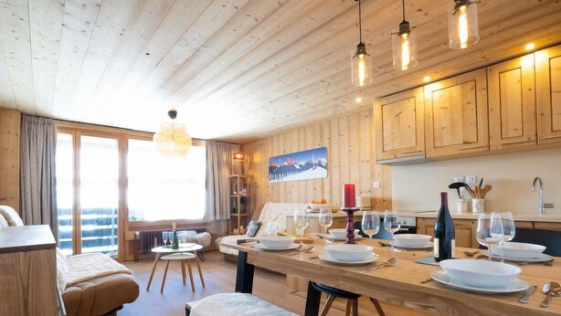 Une pièce chaleureuse et bien éclairée, avec des murs et un plafond en bois, une table à manger dressée avec des assiettes et des verres, et un banc.