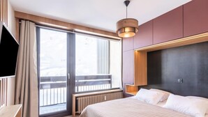 Une chambre d’hôtel moderne dotée d’un grand lit, d’une télévision, d’une porte-fenêtre coulissante donnant sur un balcon et d’un luminaire au plafond.
