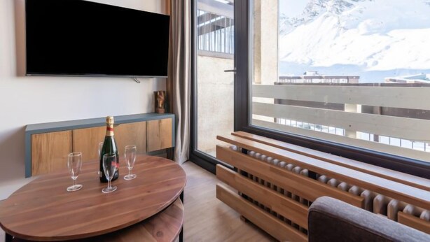 Une chambre moderne avec une table en bois, une bouteille de champagne et des verres. Un téléviseur à écran plat est fixé au mur, et une grande fenêtre offre une vue sur une montagne enneigée.