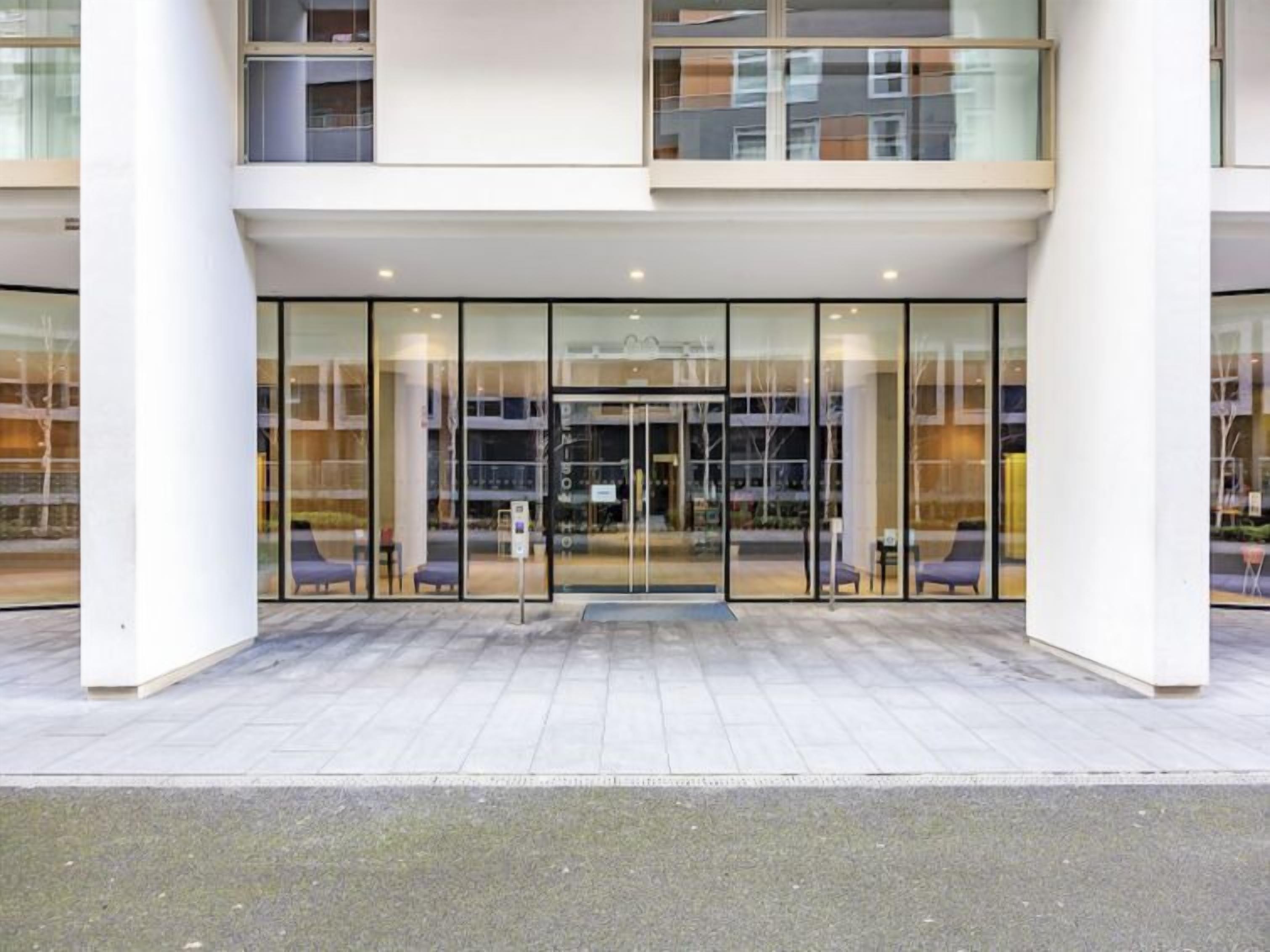 Une entrée d’hôtel moderne avec des portes en verre et un espace extérieur aménagé, abrité.