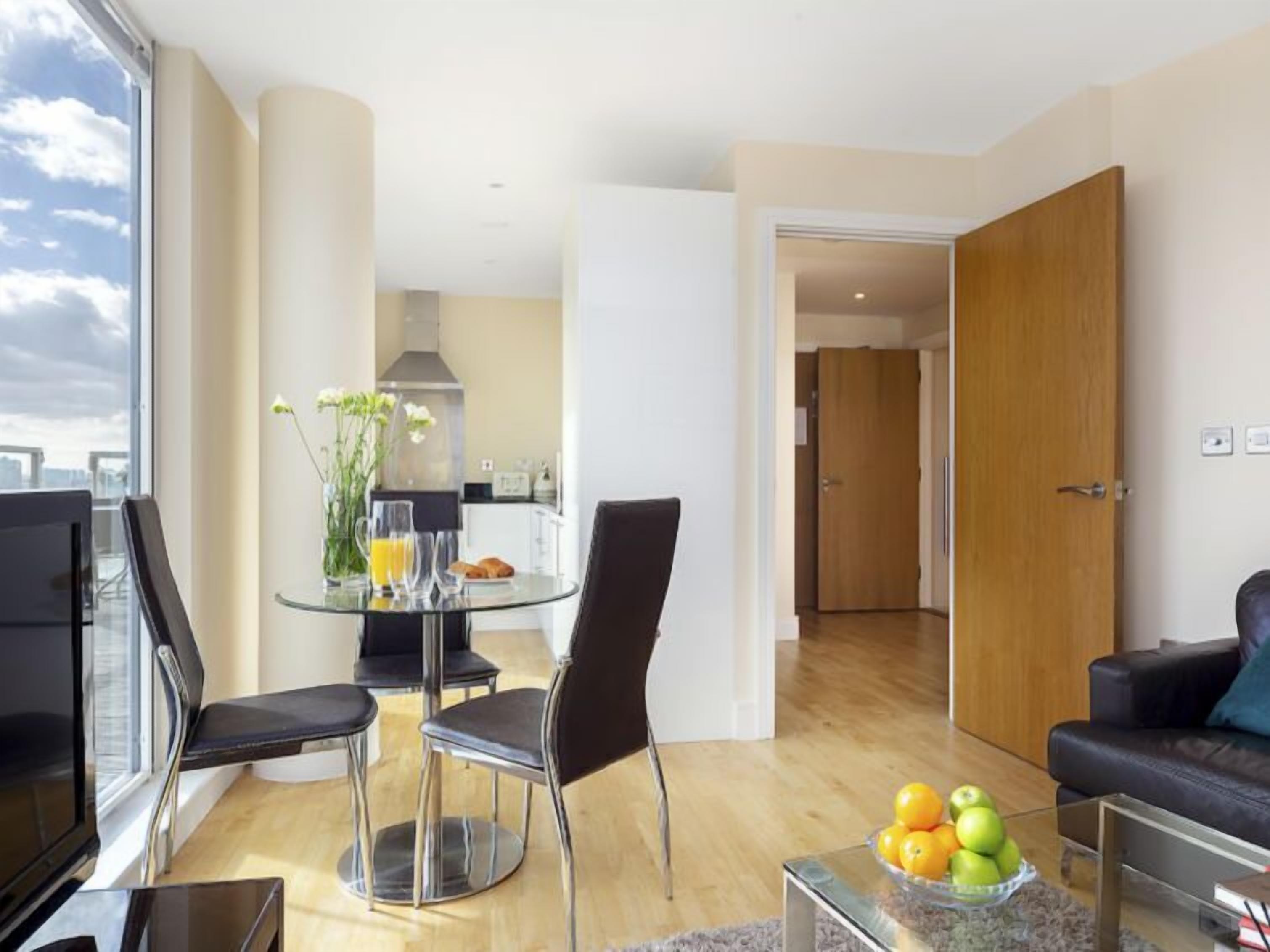 Un salon moderne avec une table à manger en verre, des chaises noires, un canapé en cuir et une table basse sur laquelle sont disposés des fruits.