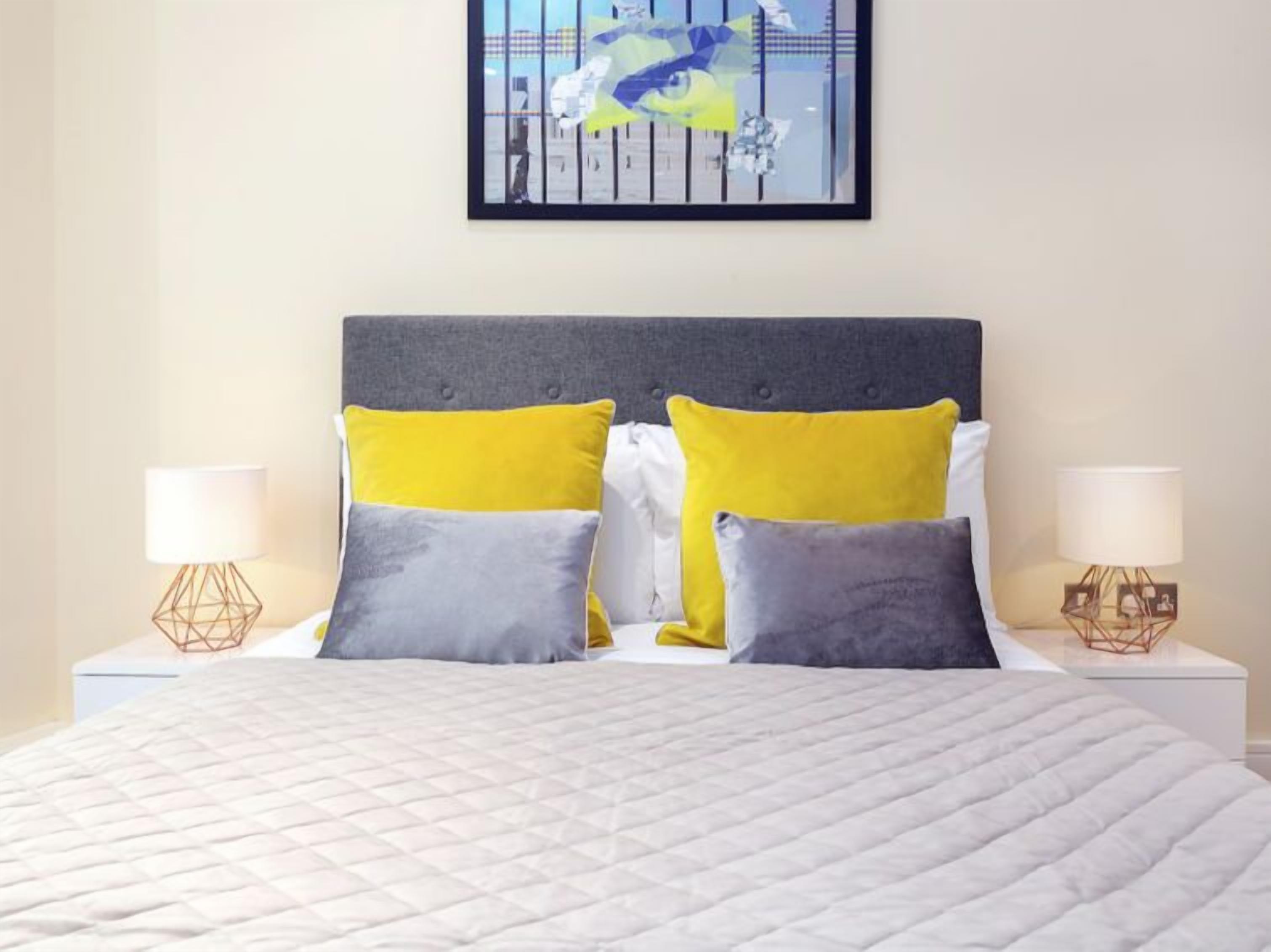 Un lit bien fait, recouvert d’une couette blanche matelassée, deux tables de chevet avec des lampes, et un tableau abstrait encadré au-dessus du lit.